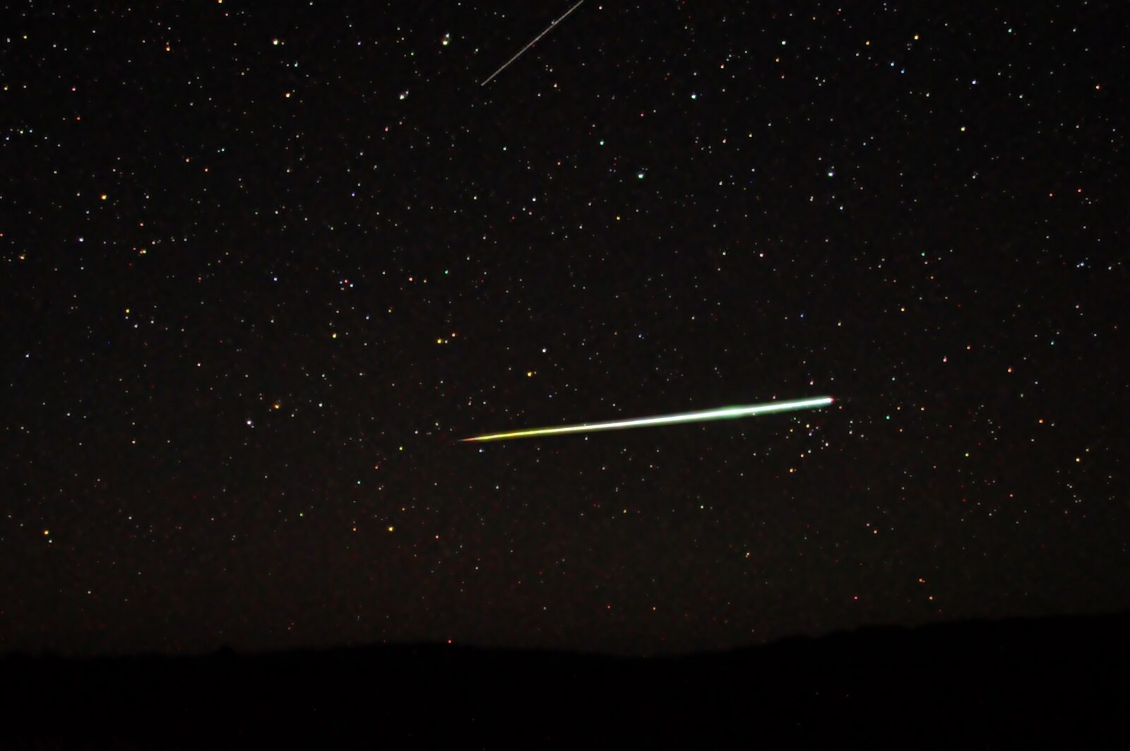 A brilliant bolide meteor streaking as a bright white-green line across a dark night sky over a desert horizon