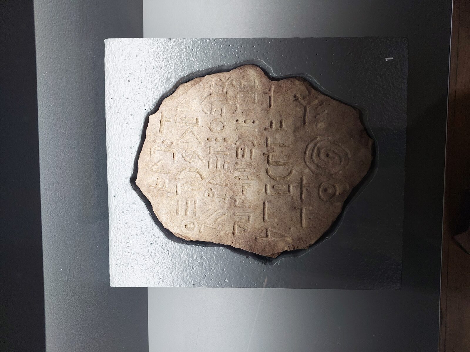 An irregular oval basalt slab mounted on a pale display block, its surface incised with rows of unfamiliar geometric signs — the Bashplemi Lake tablet.